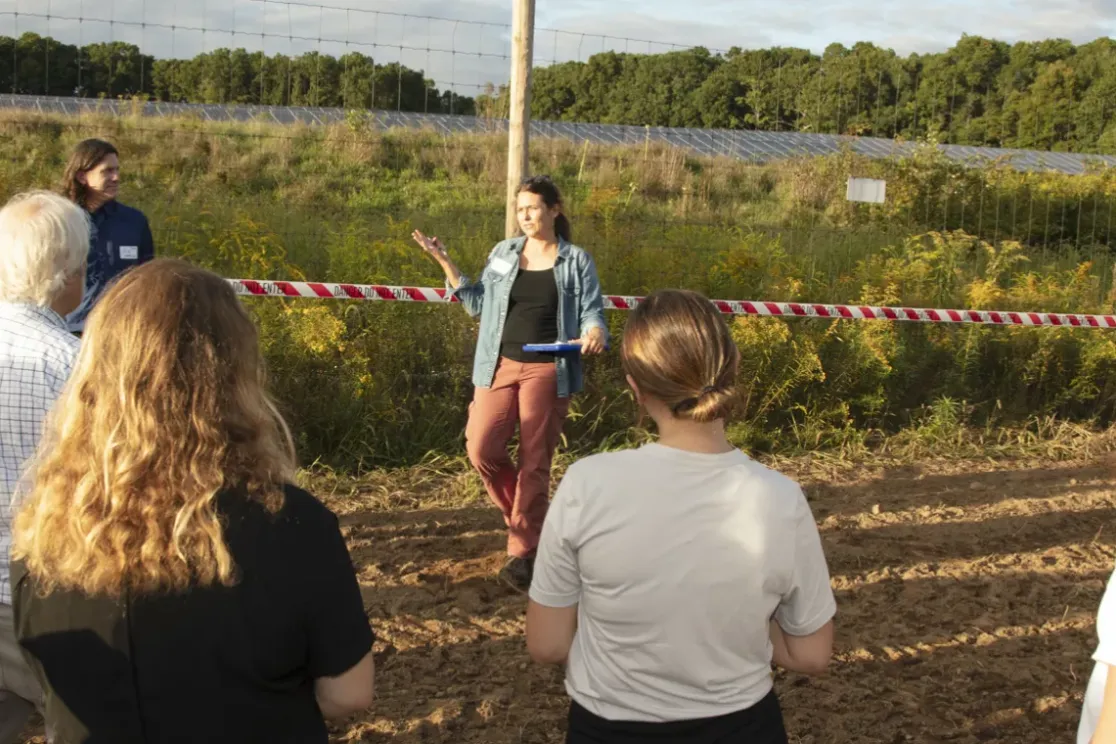 A group of people stands outdoors near a field with solar panels in the background. A woman is speaking to the group, gesturing with her hand. A red and white tape marked 'Danger Do Not Enter' separates the group from the field. Trees and greenery surround the area under a partly cloudy sky.