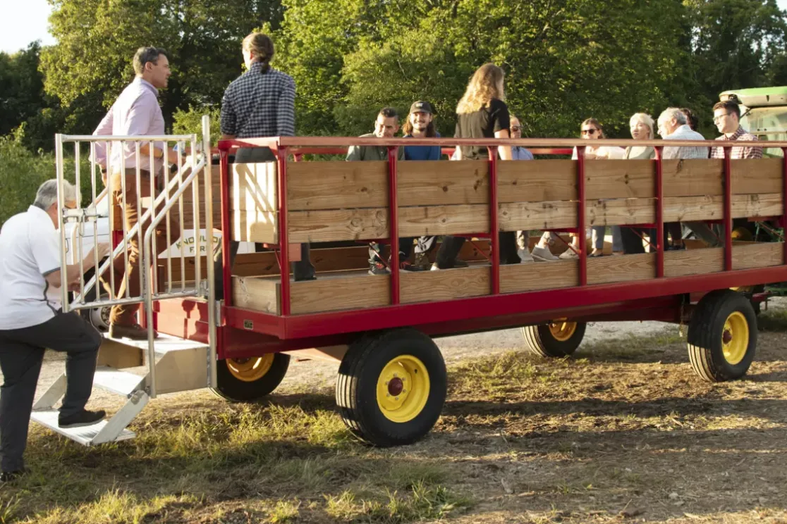 A group of people boarding a wooden wagon attached to a green tractor in a rural setting. Some are climbing stairs onto the wagon, while others are already seated. Trees and a dirt path are visible in the background.