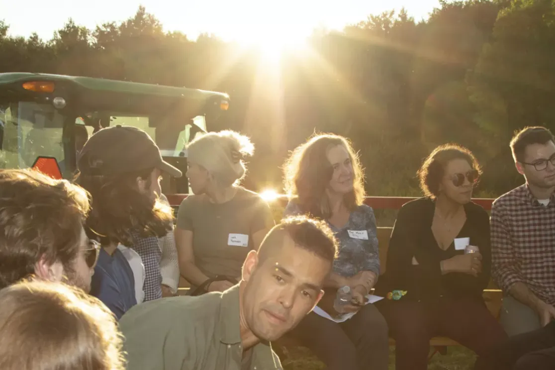 A group of people sitting outdoors in a circle, engaged in conversation. The sun is setting in the background, casting a warm glow. A green tractor is visible to the left. The group is diverse in age and gender, and some are wearing name tags.