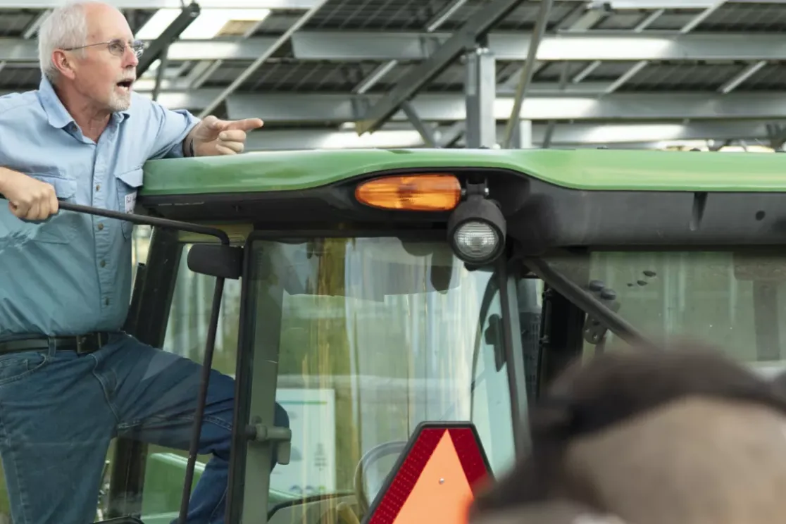 An older man in a blue shirt stands on a green tractor, pointing and speaking to a group of people. The background shows a structure with metal beams and solar panels.
