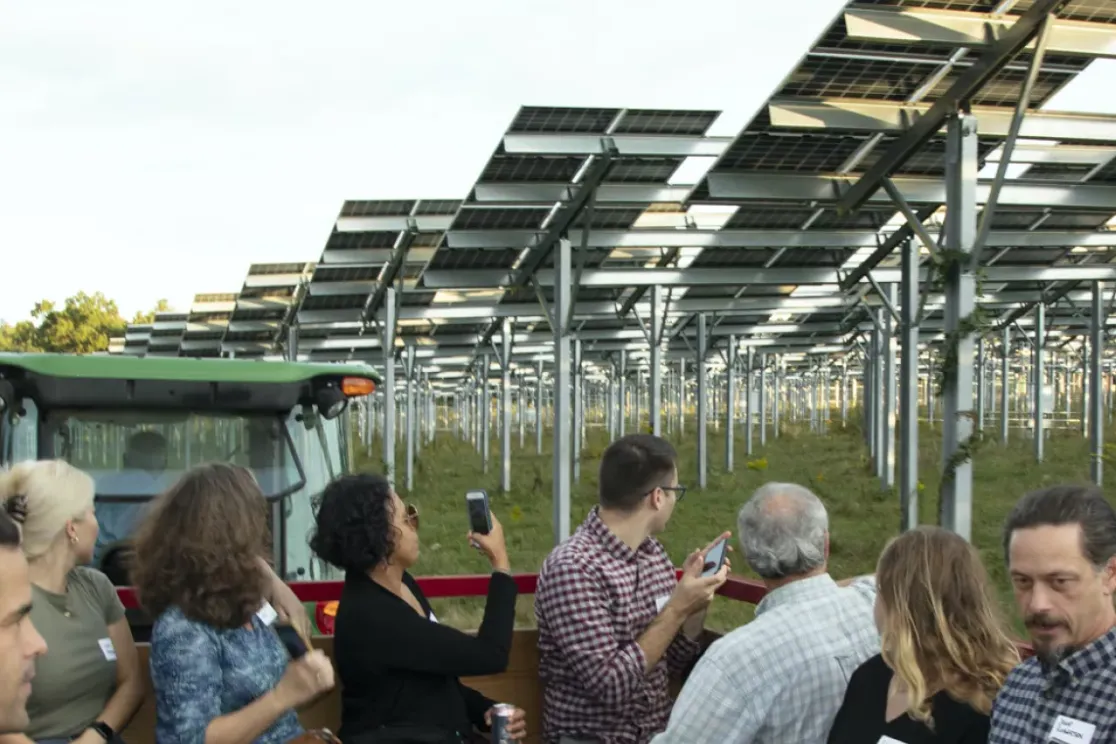 A group of people stand near a tractor, observing a field of elevated solar panels. The panels are arranged in rows, supported by metal structures. Trees are visible in the background under a clear sky.