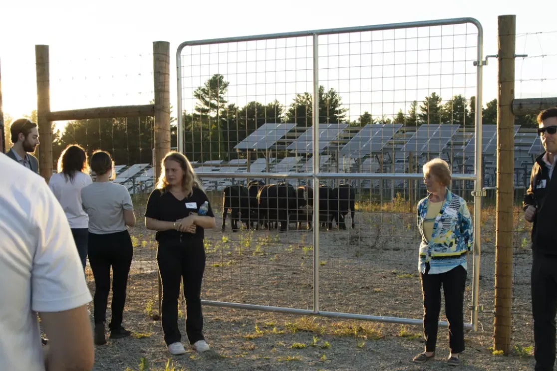 A group of people stands near a fenced area with solar panels in the background. The sun is setting, casting a warm glow. Some individuals are engaged in conversation while others observe the surroundings.