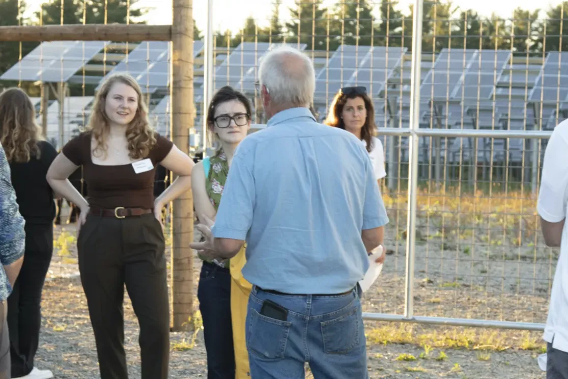A group of people stands outside near solar panels, engaged in conversation. The setting is sunny, and they are casually dressed. The background features a fence and rows of solar panels.