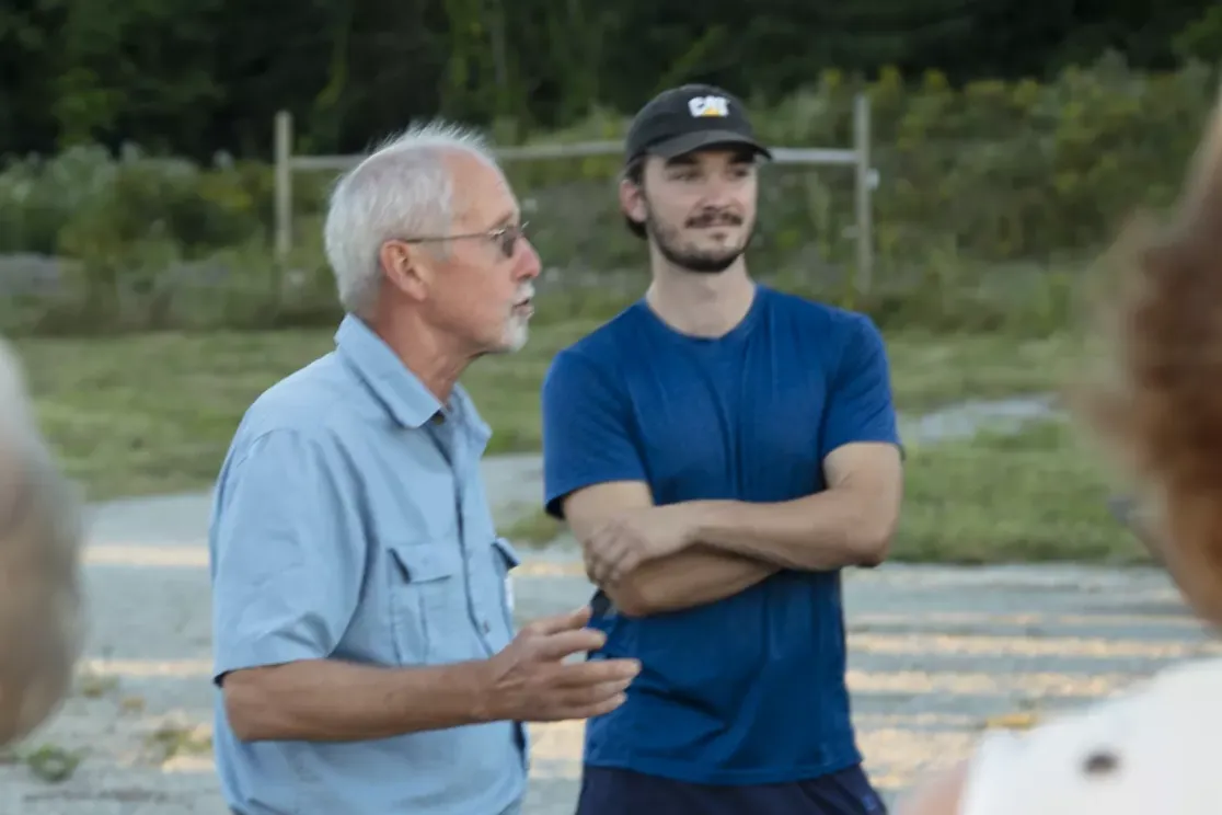 A group of people gathered outdoors in a conversation. An older man in a light blue shirt is speaking, while a younger man in a blue T-shirt with crossed arms listens. Others are partially visible, with trees and grass in the background.