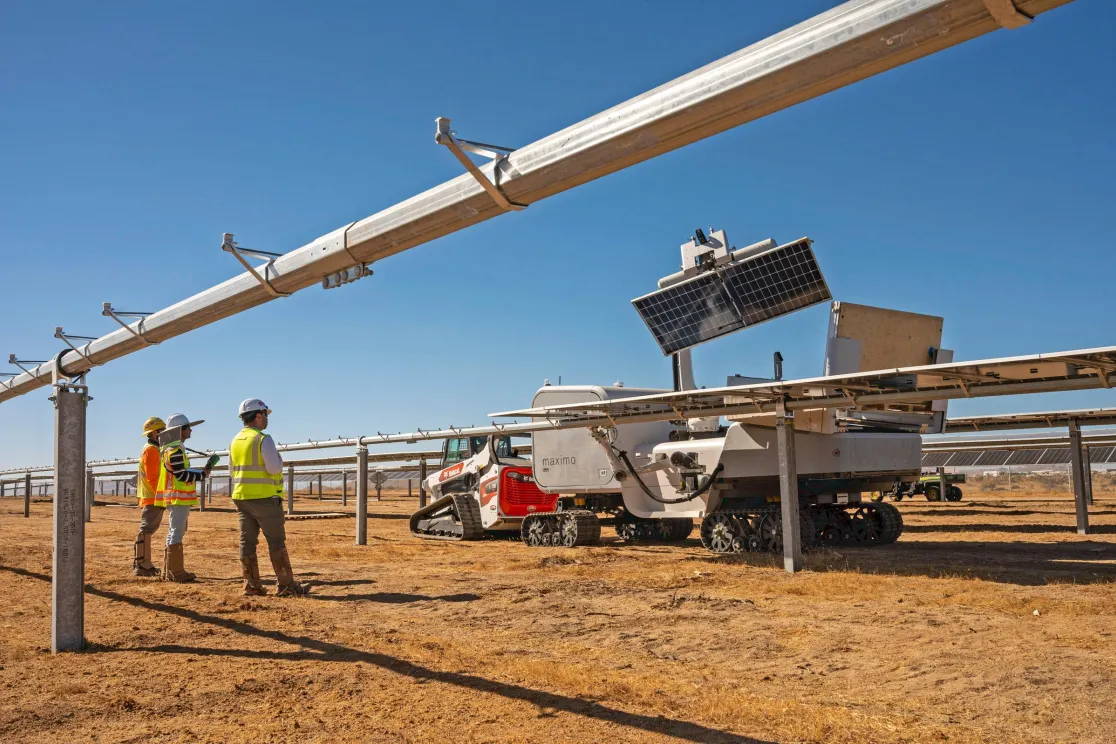 Three workers in safety gear stand near a solar panel installation site with a robotic vehicle on a sunny day.