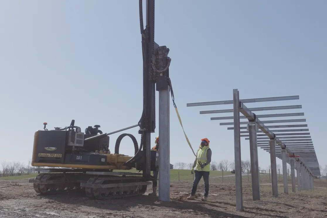 A construction worker in a safety vest and helmet stands next to a pile driver machine installing metal posts in a field. The posts form a long row extending into the distance. The sky is clear and blue.