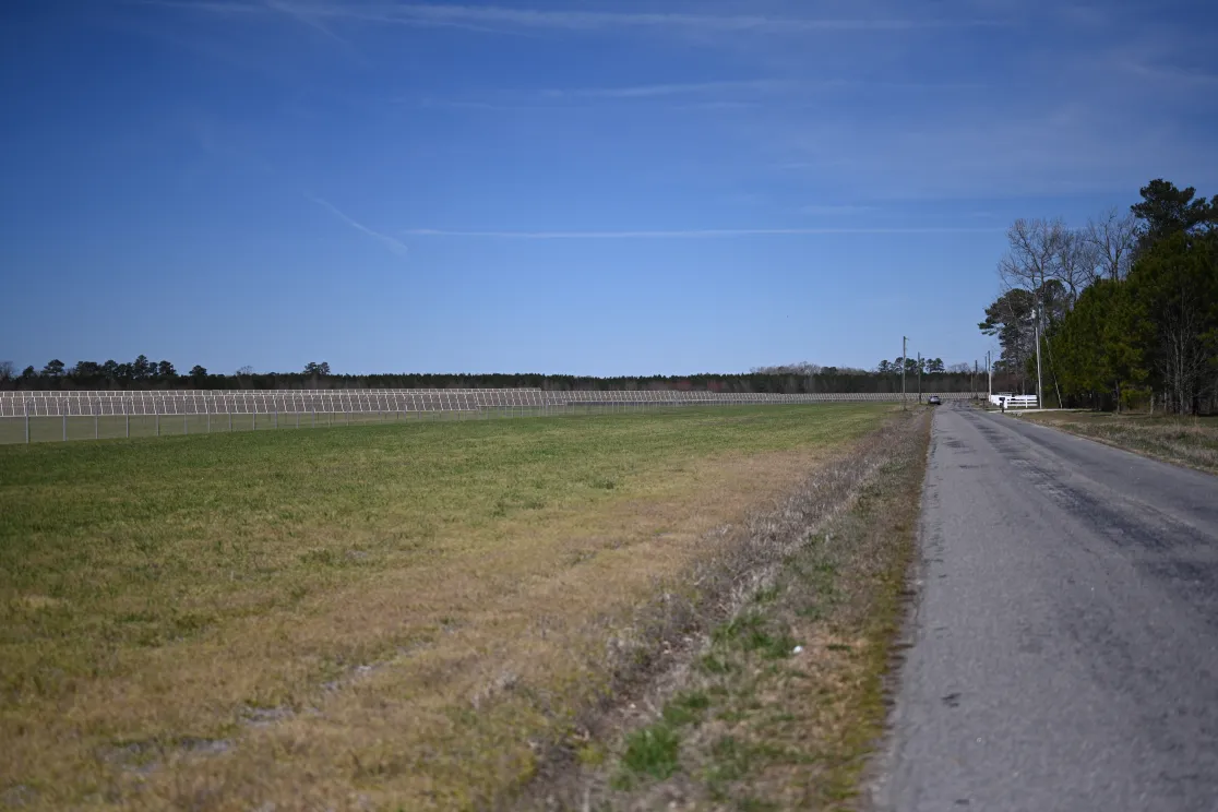 A rural landscape featuring a narrow paved road on the right and a large grassy field on the left. A row of solar panels is visible in the distance, parallel to the road. The sky is clear and blue, with some trees lining the horizon.