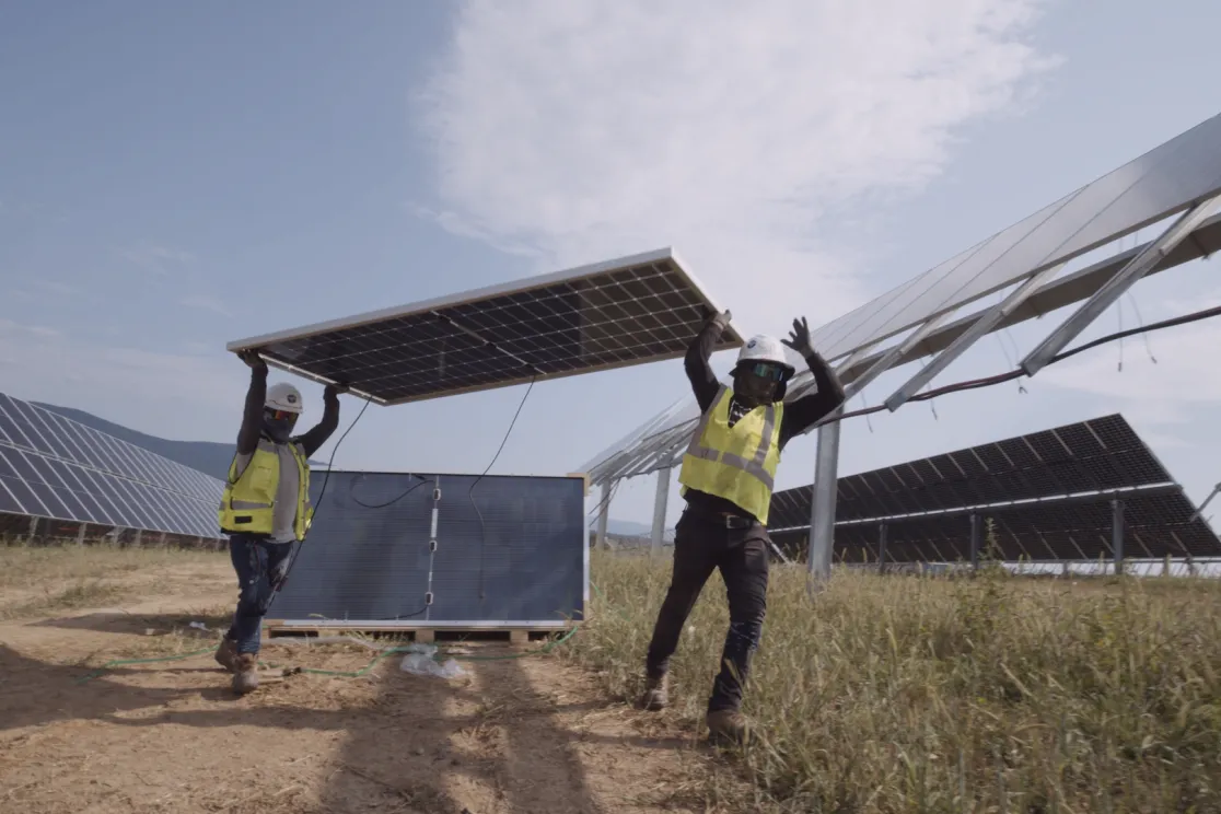 Two workers in safety gear and helmets carry a large solar panel through a solar farm. Rows of solar panels are visible in the background under a partly cloudy sky.