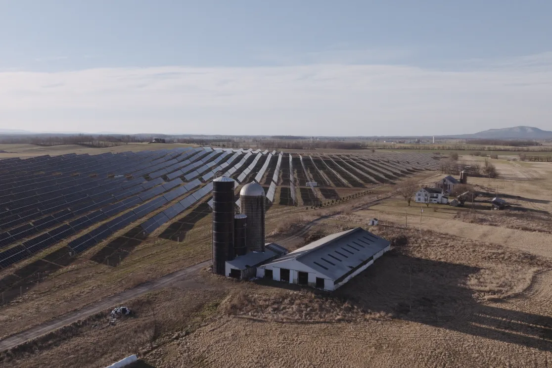 Aerial view of a large solar farm with rows of solar panels stretching across a vast field. In the foreground, there is a barn and silos. The landscape is rural with distant mountains under a clear sky.