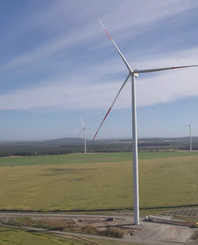 A landscape with several wind turbines in a vast field under a blue sky with light clouds. The turbines are tall with large blades, and there are vehicles and equipment at the base of the nearest turbine.