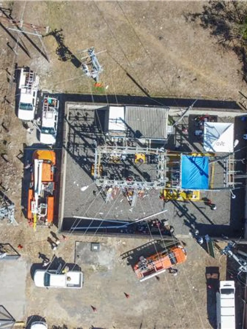 Aerial view of a power substation with multiple vehicles and equipment around it. The substation is surrounded by a fence and located near a road. The area is dry with some vegetation nearby.