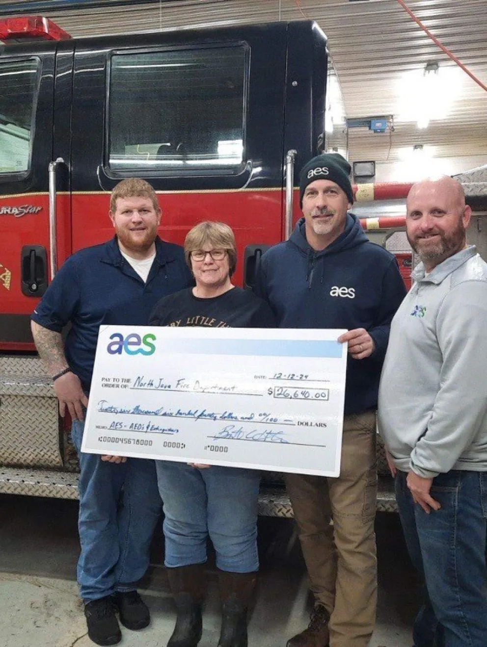 Four people stand in front of a North Java Fire Company truck holding a large check for $26,640 from AES. The group is smiling and appears to be inside a fire station.
