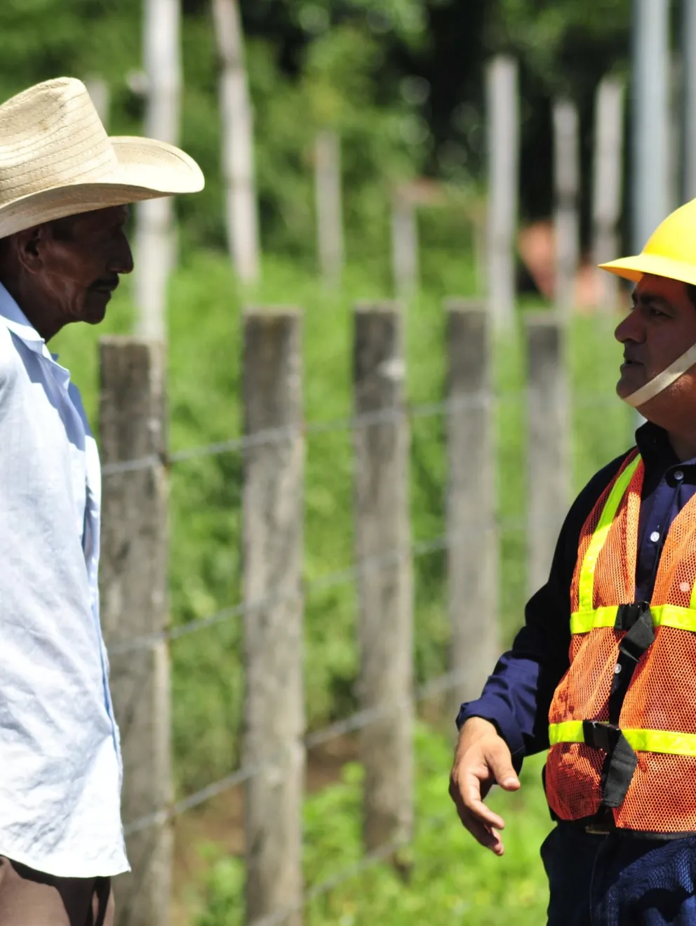 Two men having a conversation outdoors. One wears a straw hat and light shirt, the other a yellow hard hat and reflective vest. They stand by a fence with greenery in the background.
