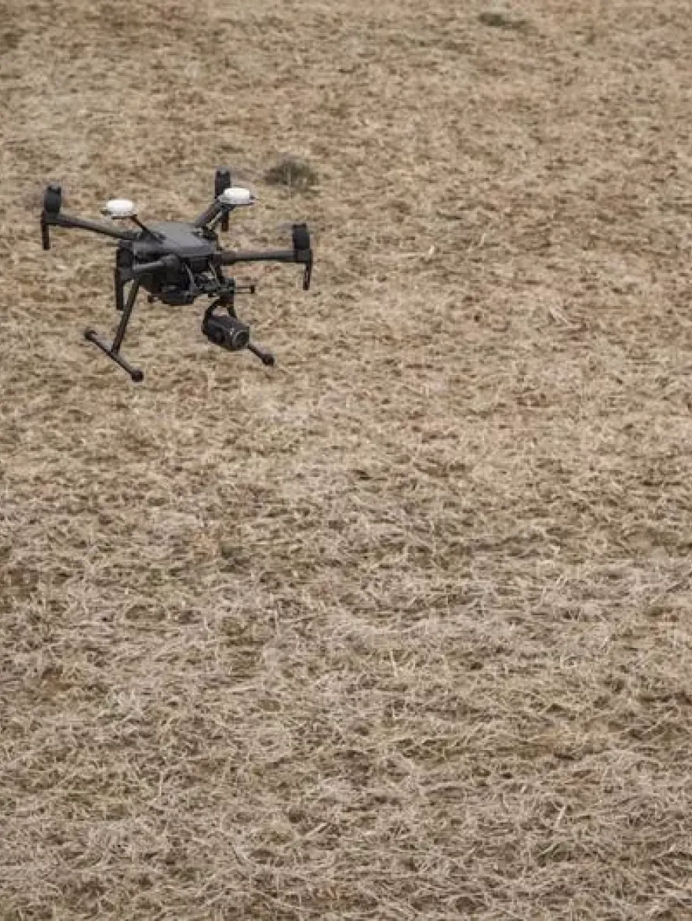 Two people in yellow safety jackets and white helmets stand in a field, operating a drone that is flying nearby.