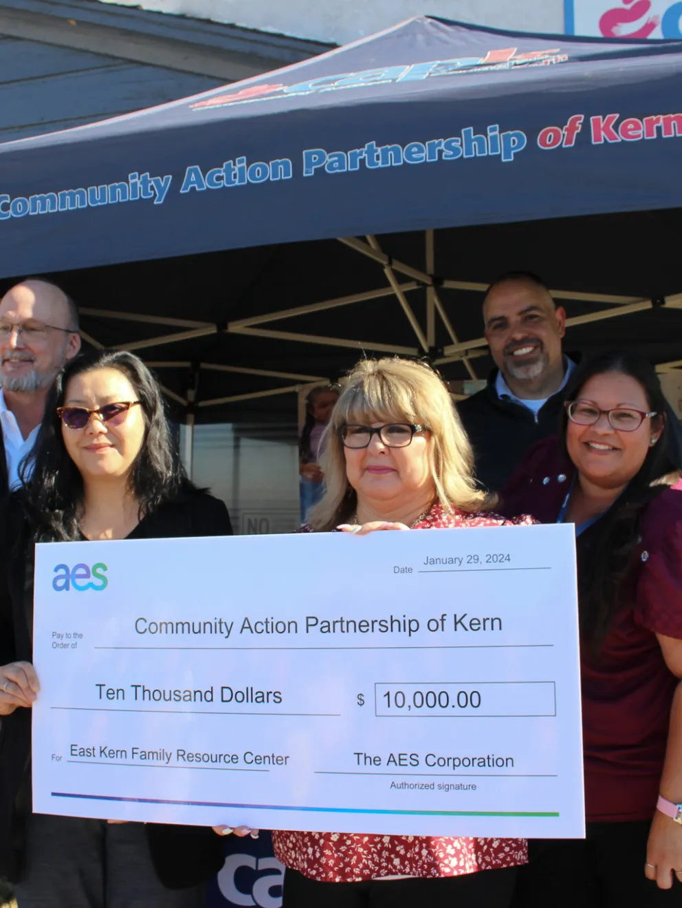 A group of five people stands under a canopy labeled 'Community Action Partnership of Kern.' They are holding a large check for ten thousand dollars from AES Corporation to the East Kern Family Resource Center. Two banners on either side display information about combating food insecurity and empowering youth and families.