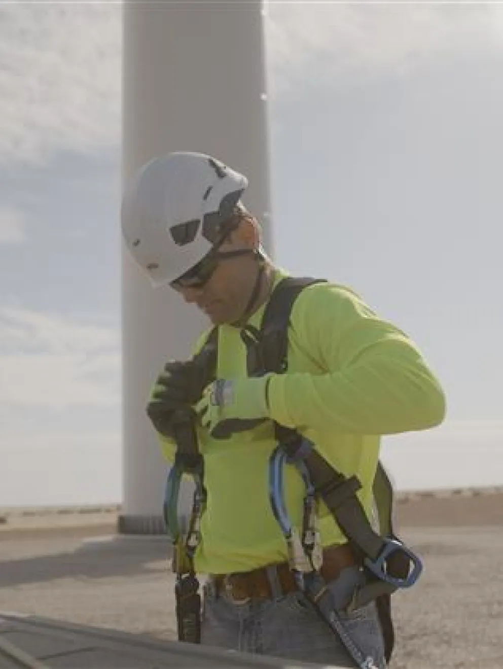 A person wearing a bright yellow safety jacket and helmet adjusts their harness near a wind turbine. The scene is outdoors with a clear sky.