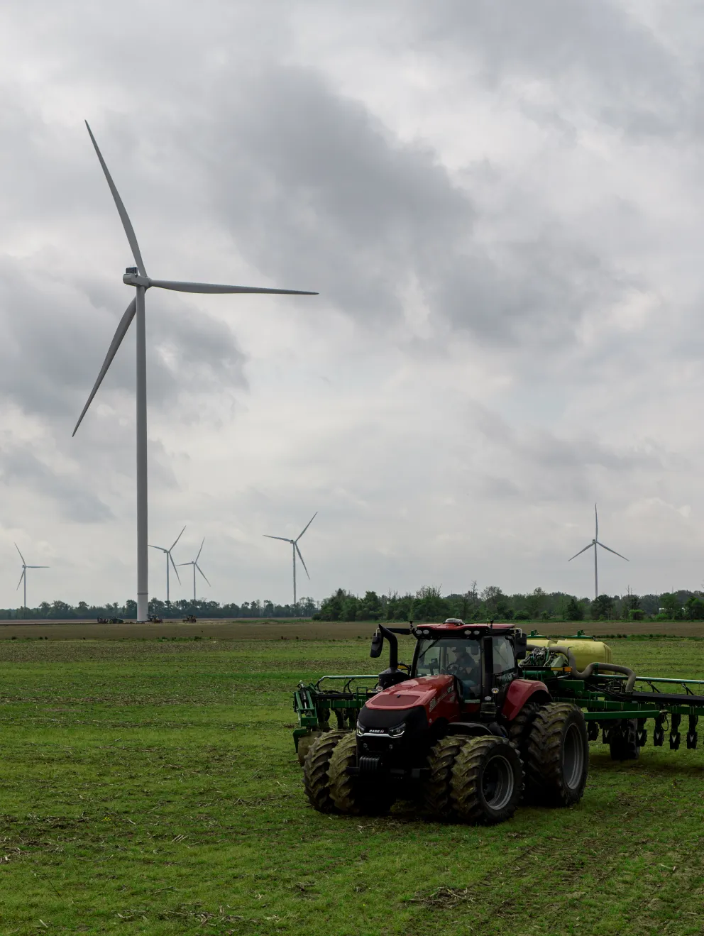 A red tractor with large tires is parked on a green field under a cloudy sky. In the background, several wind turbines are visible across the landscape, with one prominently in the foreground. Trees line the horizon.