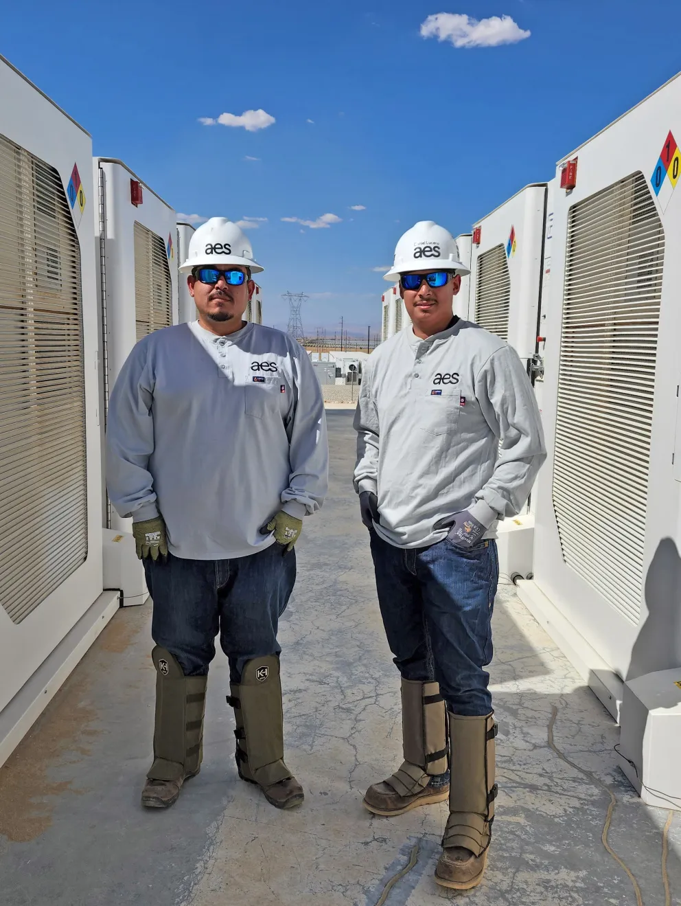 Two workers wearing AES-branded helmets and protective gear stand between large white industrial units under a clear blue sky.
