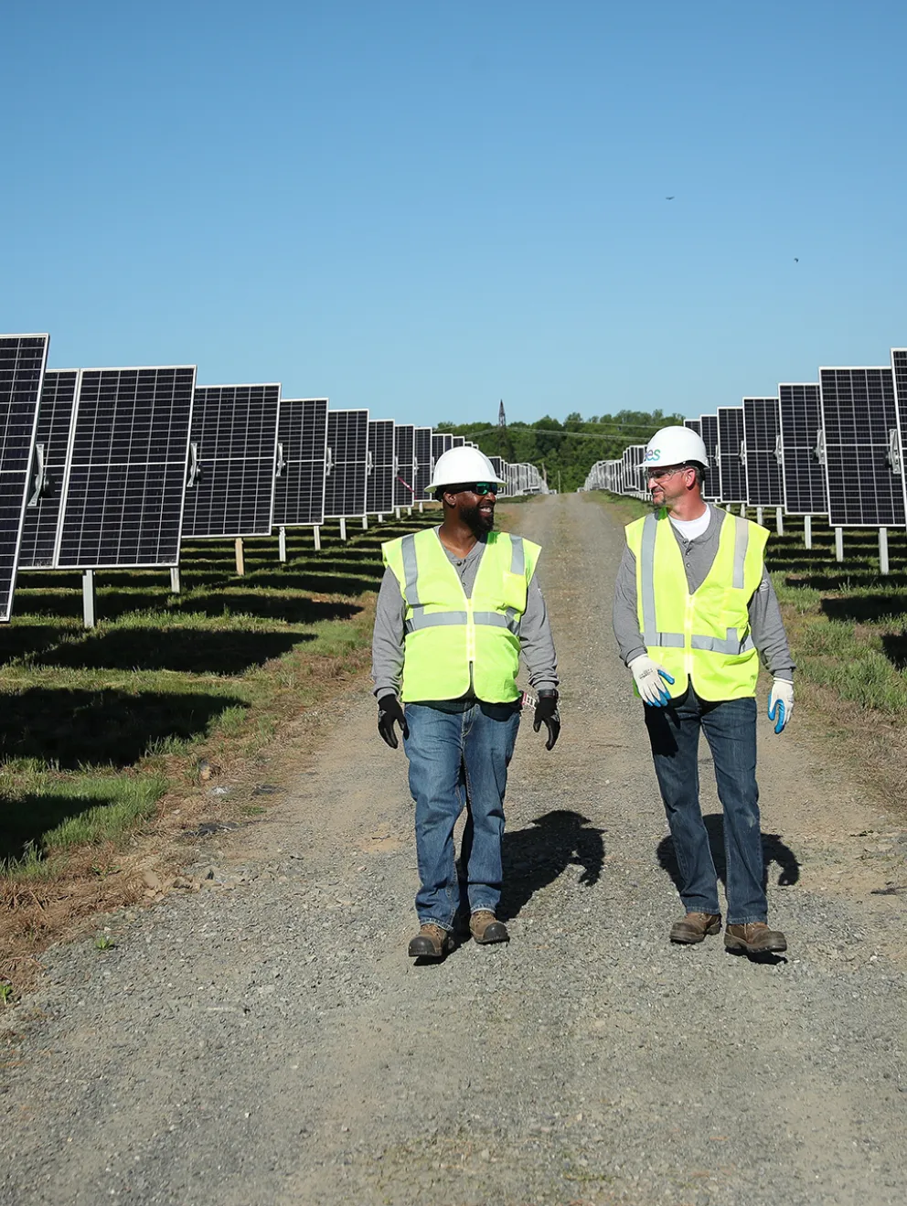 Two workers in safety vests and helmets walk along a path between rows of solar panels under a clear blue sky.