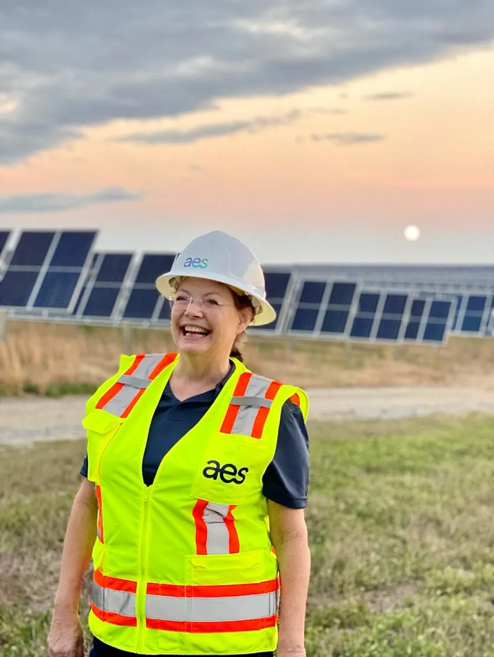 A smiling person wearing a white hard hat and a bright yellow safety vest with 'aes' logo stands in front of solar panels at sunset, with a grassy field and sky in the background.