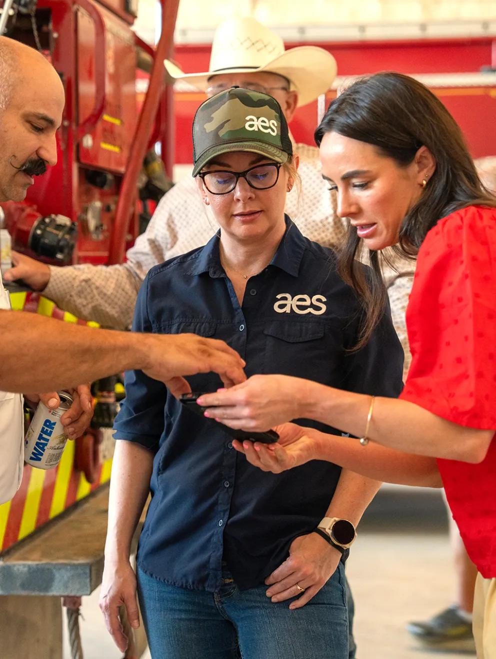 Three people standing in front of a fire truck, collaborating and looking at a smartphone. One person wears a white uniform, another wears a blue shirt and cap with 'aes' logo, and the third person wears a red shirt. The setting appears to be a fire station.
