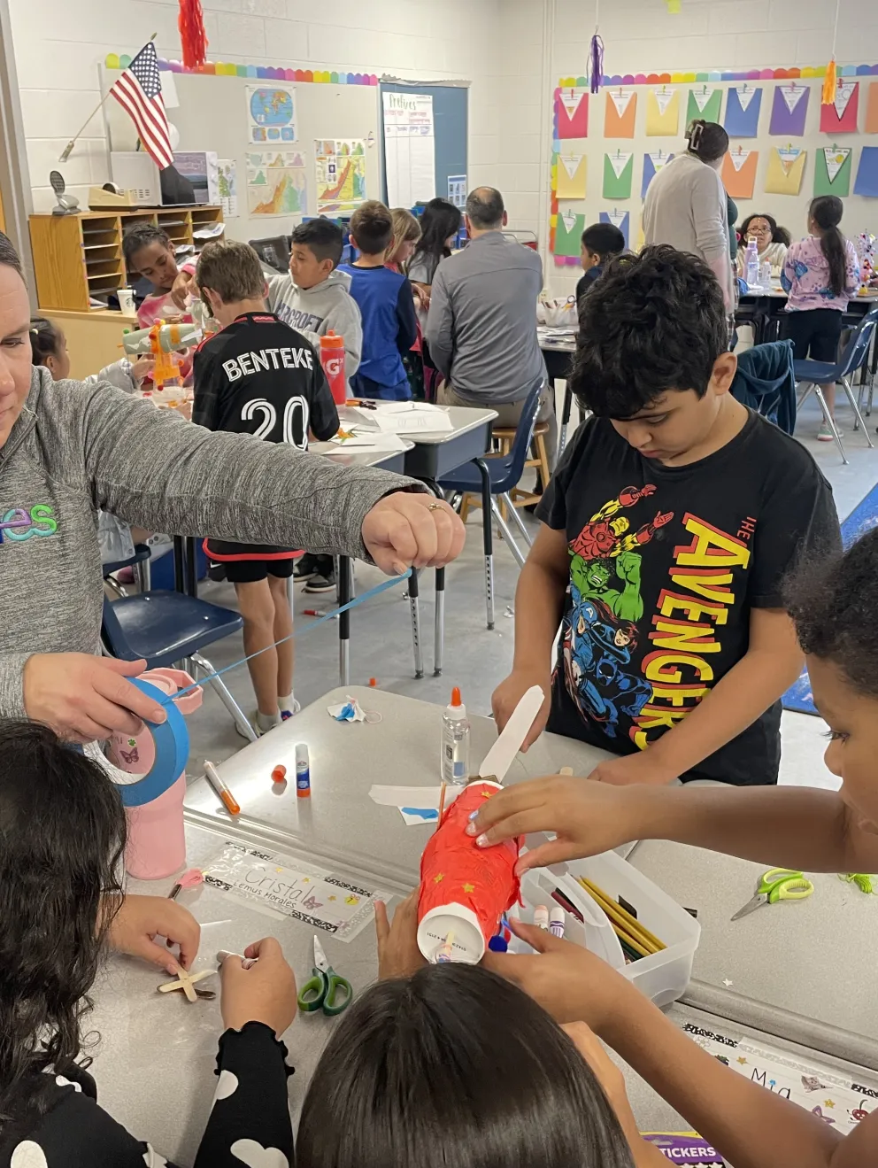 A teacher assists children with a craft project in a classroom. The students are engaged in creating art using various materials, including tape and paper. The classroom is decorated with colorful artwork and an American flag. Other students and teachers are visible in the background working on similar projects.