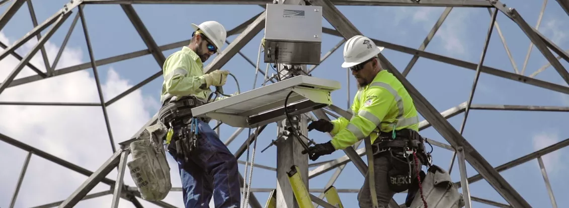 Two utility workers in safety gear and helmets are installing equipment on a metal tower. They are using ladders and are surrounded by a framework of steel beams against a blue sky.
