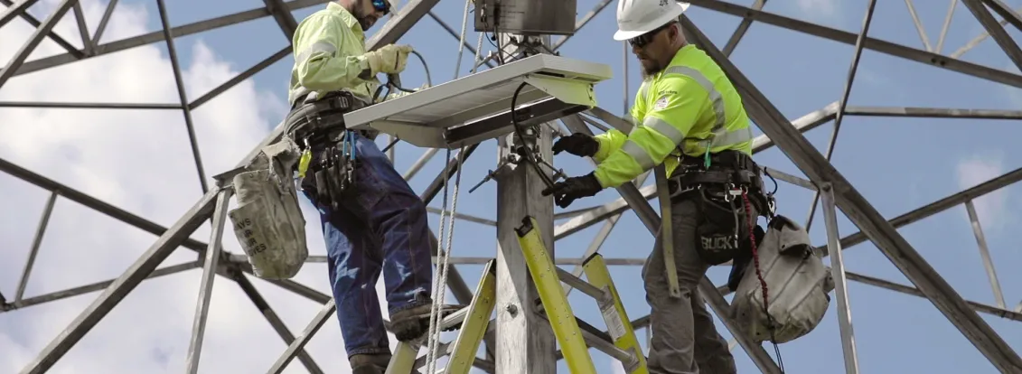 Two workers in safety gear install equipment on a metal tower against a blue sky.