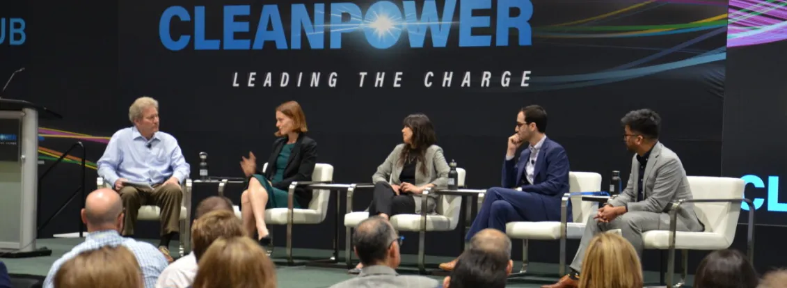 A panel of five people seated on a stage at a CleanPower event, with an audience in the foreground. The backdrop displays the words 'CleanPower Leading the Charge.'