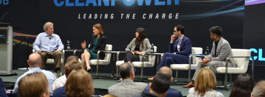 Panel discussion at Clean Power event with five speakers seated on stage, engaging with an audience. The backdrop displays the words 'Clean Power Leading the Charge'.