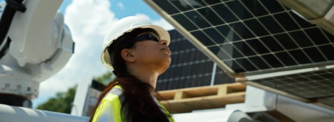 A person wearing a hard hat and safety vest inspects solar panels under a clear blue sky.