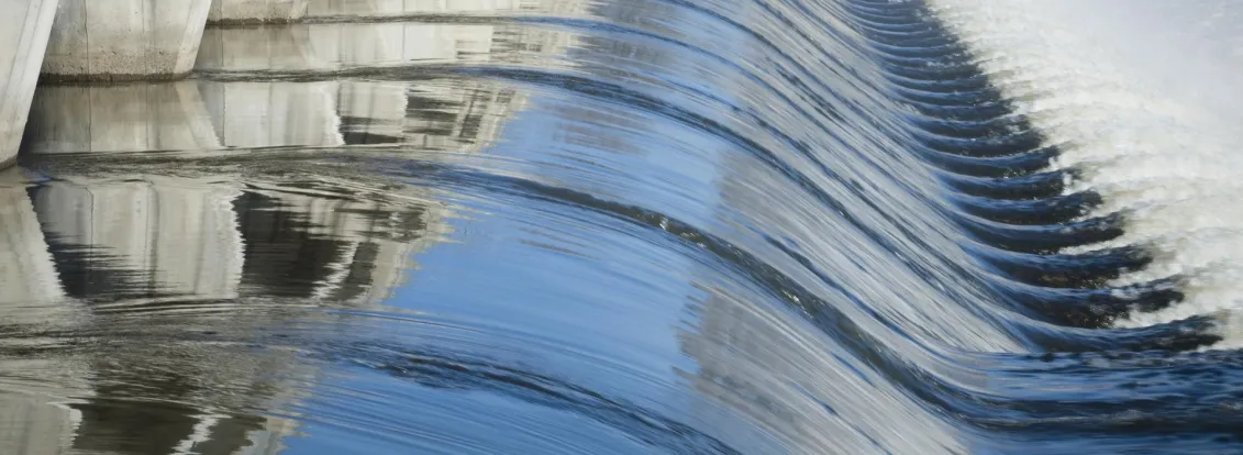A large dam with water cascading over its edge, creating smooth, wavy patterns. The concrete structure is reflected in the water, and mist rises from the flowing water on the right.