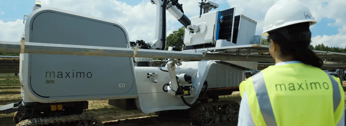 A person wearing a safety vest and hard hat observes a solar panel installation robot labeled 'maximo' on a sunny day.