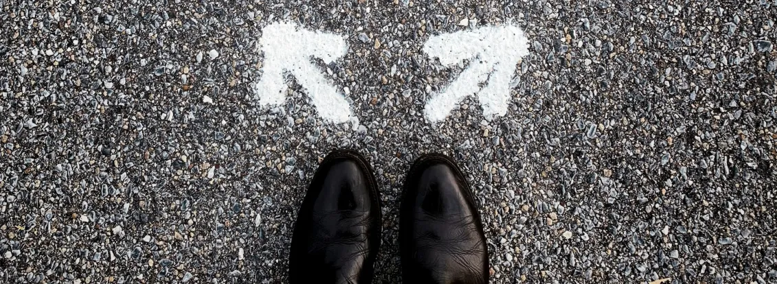 A person wearing black shoes stands on a gravel road in front of two white arrows painted on the ground, pointing in different directions, symbolizing a choice or decision.