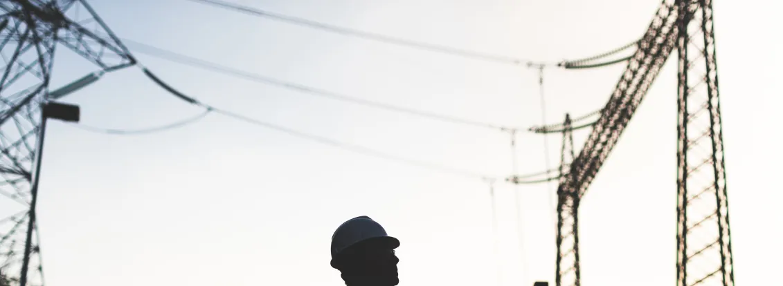 Silhouette of an electrical worker wearing a hard hat standing in front of high voltage transmission towers at sunset.