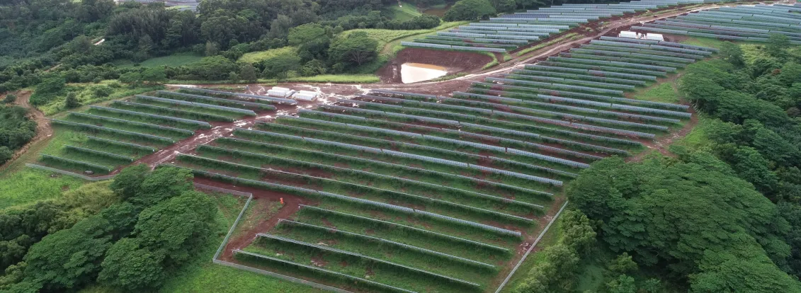 Aerial view of a large solar farm with rows of solar panels on a green landscape surrounded by trees and hills.