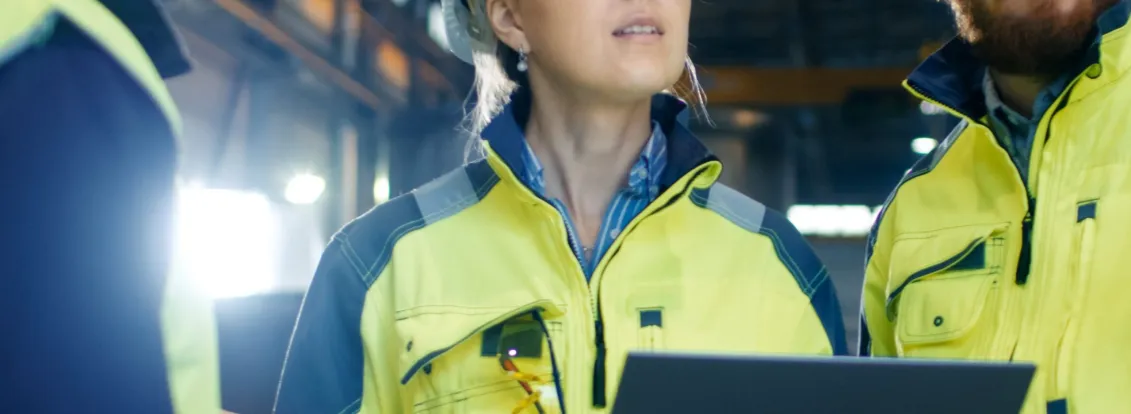 Three construction workers wearing safety helmets and high-visibility jackets stand inside a large industrial facility. One holds a laptop, and they appear to be discussing or inspecting something above.