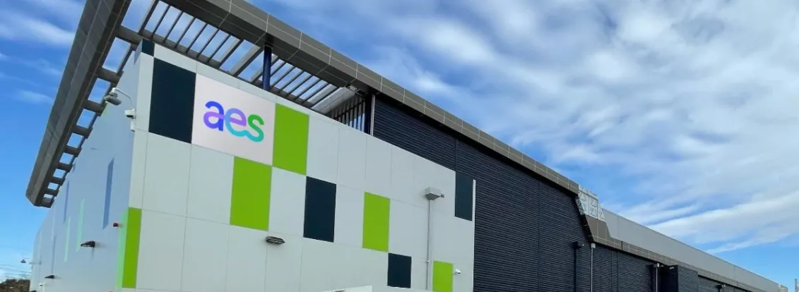 A modern industrial building with a geometric design and the letters 'aes' on the facade. The building features a mix of white, green, and dark panels, with several electrical units in front. The sky is partly cloudy.