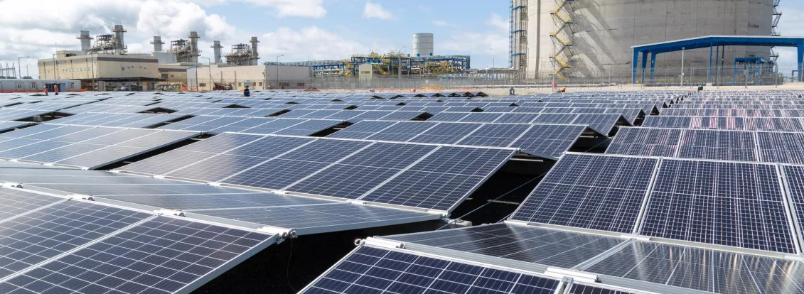 A large array of solar panels under a blue sky, with an industrial facility and a large cylindrical structure in the background.