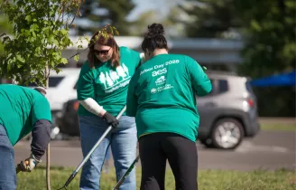 A group of people wearing green shirts participate in a tree planting event, with one person holding a shovel near a young tree. A car is parked in the background.