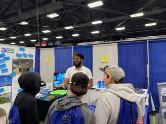 A man in a white polo shirt engages with three young people at a booth during a career fair. The booth displays information about renewable energy, with a banner featuring wind turbines. The attendees wear backpacks with 'Career Fair' written on them.