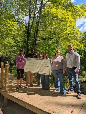 Six people standing on a wooden platform in a forest, holding a large check from AES. The background shows green trees and a clear blue sky.