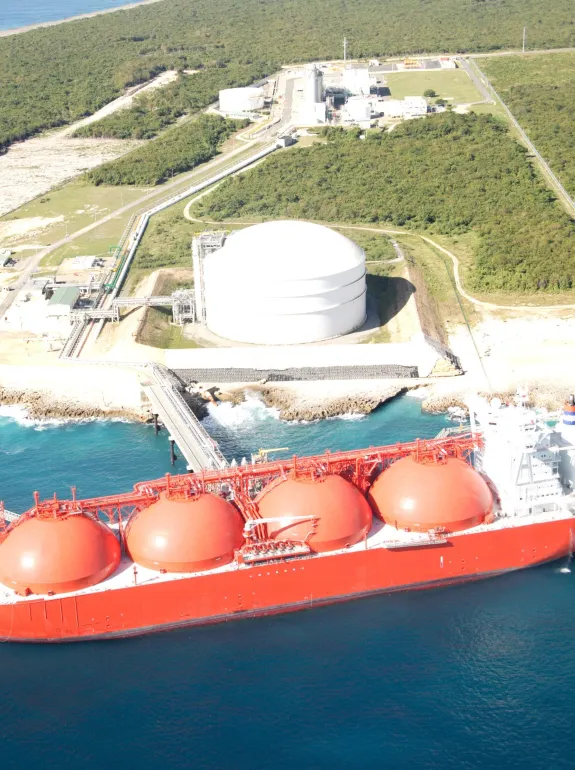 Aerial view of a large red LNG tanker docked at a coastal terminal with spherical storage tanks and surrounding greenery.