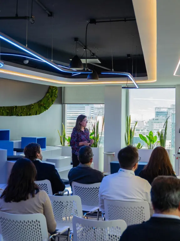 A woman presents in a modern conference room with plants and large windows. Attendees sit in white chairs facing a screen displaying a presentation titled 'Acuerdo de París'.