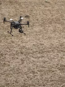 Two people in high-visibility jackets and helmets stand in a field, operating a drone that is flying nearby.
