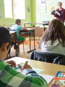 A classroom with students sitting at desks working on assignments. A teacher stands near a whiteboard holding a book. The classroom is brightly lit with green walls and posters. An English textbook is visible on one of the desks.