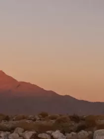 Two wind turbines stand in a rocky desert landscape at sunset, with a mountain range in the background illuminated by a warm pink glow.
