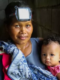 A woman wearing a headlamp holds a baby wrapped in a blue patterned blanket. The woman looks at the camera with a slight smile, while the baby looks curiously. The background is dimly lit, highlighting the subjects.