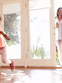 A young girl joyfully runs across a sunlit living room with wooden floors. Behind her, a family enters through open double doors, smiling and holding a baby.