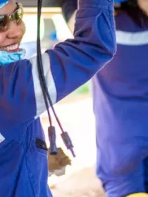 A person wearing a blue hard hat, safety glasses, and a blue uniform with reflective stripes is smiling while working on an electrical panel. They are outdoors, with another person in similar attire working nearby.